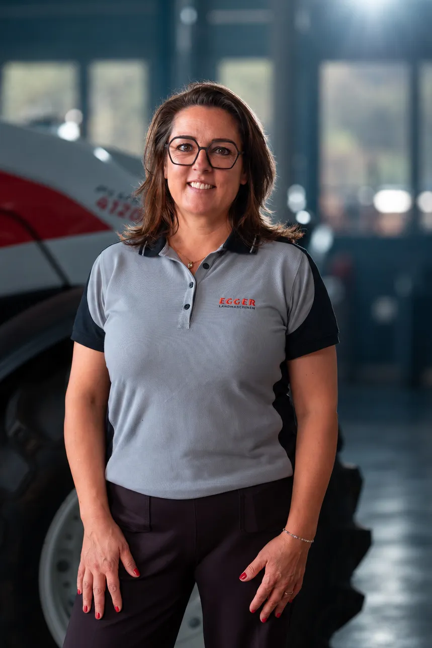 Corinne Egger mit Brille im grauen Egger-Polohemd, hinter ihr ein roter Traktor.