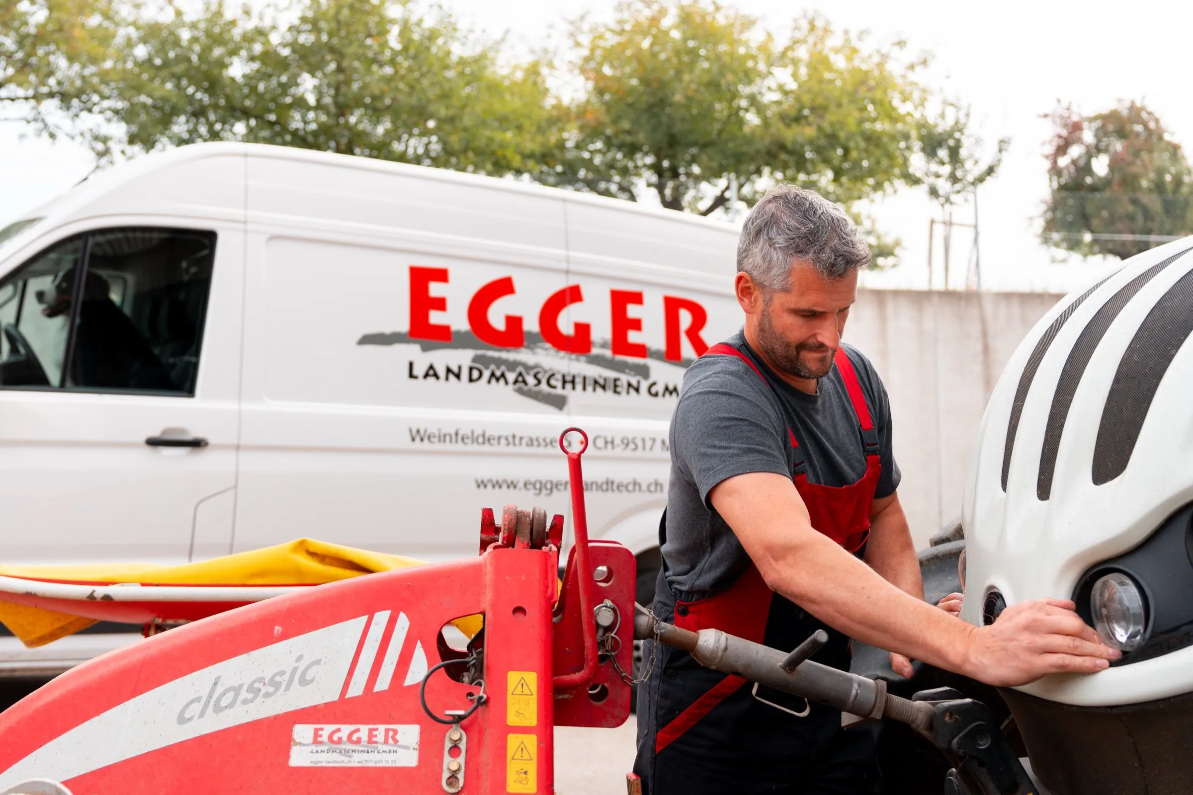 Ein Mechaniker arbeitet am Scheinwerfer eines Traktors, dahinter stehen ein Mähwerk und der beschriftete Servicebus von Egger.